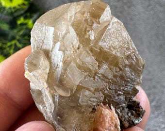May include: A raw, natural crystal cluster with a light tan color and a rough, textured surface. The crystal has multiple facets and a small, reddish-brown inclusion at the base. The crystal is held in a hand.