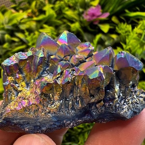 May include: A cluster of iridescent aura quartz crystals. The crystals display a spectrum of colors, including blues, purples, and golds, with a textured, rough base. The crystals are held in a hand, with green foliage in the background.
