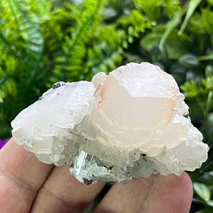 Puede incluir: Un grupo de cristales de calcita transparentes, blancos y rosados con un toque de verde. Los cristales están dispuestos en una formación en capas, con el cristal más grande en la parte superior. Los cristales tienen una textura natural y rugosa.