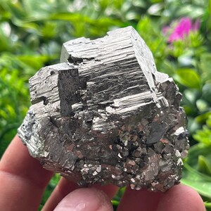 Puede incluir: Un gran cristal de pirita de color plateado con forma cúbica. El cristal tiene una superficie rugosa y texturizada y está compuesto por muchos cristales más pequeños.