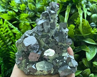 May include: A cluster of dark gray, metallic, faceted crystals with a rough, textured surface. The crystals are arranged in a pyramid shape.