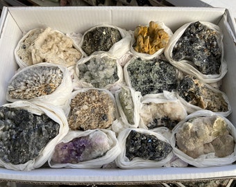 May include: A collection of various crystals and minerals, including amethyst, quartz, and pyrite, are displayed in a white box. The crystals are wrapped in white tissue paper.
