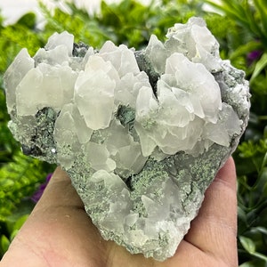 Puede incluir: Un cúmulo de cristales de calcita blancos sobre una matriz gris. Los cristales son translúcidos y tienen un ligero tono verde. La matriz es rugosa y texturizada.