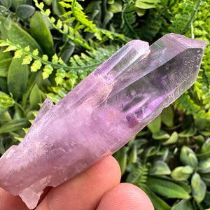 Pode incluir: Uma ponta de cristal de ametista roxo-claro translúcido com uma forma hexagonal alongada natural. O cristal apresenta uma superfície lisa e facetada com inclusões internas visíveis e uma terminação pontiaguda na extremidade superior.