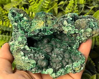 May include: A large, green malachite specimen with a rough, textured surface. The malachite has a deep green color with some lighter green accents. The specimen has a unique, cave-like shape with a hollow center.