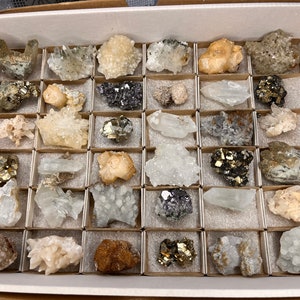 May include: A collection of 36 different types of crystals and minerals displayed in a cardboard box with individual compartments. The crystals are various colors, including white, clear, brown, black, and gold.