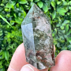 Pode incluir: Uma ponta de cristal clara e facetada com inclusões verdes, castanhas e brancas. O cristal é segurado em uma mão, com um fundo verde desfocado de folhagem. O cristal tem uma superfície polida e lisa.