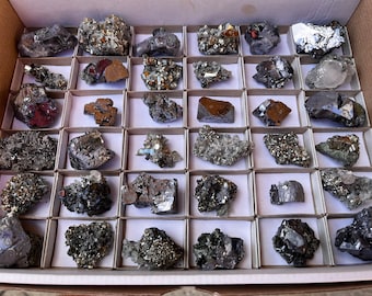 May include: A collection of 25 gray and silver mineral specimens in a white box with individual compartments. The minerals are arranged in rows and columns, and each specimen is displayed on a white background.