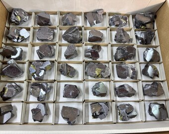 May include: A cardboard box filled with individual small cardboard boxes, each containing a dark, irregularly shaped mineral specimen. Some specimens have metallic gold and silver colored inclusions. The minerals are arranged in a grid pattern.