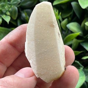 Puede incluir: Un cristal blanco en forma de pera con una superficie rugosa y texturizada. El cristal tiene un color blanco ligeramente apagado y está cubierto de pequeños puntos blancos.