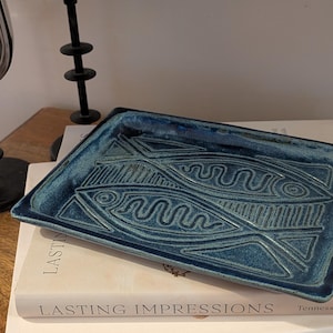 May include: A rectangular ceramic tray with a deep blue, textured glaze. The surface features an embossed design of two stylized fish with geometric patterns and wavy lines. The tray has slightly raised edges and a glossy, mottled finish.