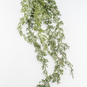 May include: A cascading, artificial evergreen plant with small, needle-like leaves. The plant is a vibrant green color and is set against a plain white background. This decorative plant is suitable for home decor.