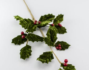 May include: A decorative holly branch featuring green velvet-textured leaves and small, glossy red berries attached to a thin, light brown stem. The artificial botanical piece displays multiple clusters of leaves and berries arranged along the branch.