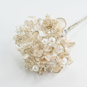 May include: A decorative floral bouquet with cream-colored petals, accented with gold glitter and faux pearls. The arrangement includes clear crystal beads and a silver stem, ideal for weddings or special events.