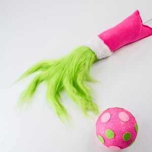 May include: A whimsical Grinch-themed plush toy set. The set includes a pink arm with green fur fingers, a white cuff, and a pink ball with green and pink polka dots. Perfect for holiday decor.