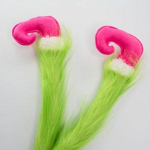 May include: Two whimsical Christmas decorations featuring bright pink curled hats with white trim, atop long, fluffy green faux fur legs. The festive accents are set against a plain white background.