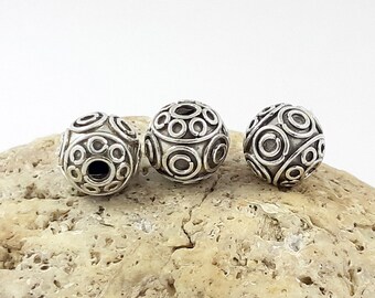 May include: Three silver-colored, spherical beads with an intricate design of concentric circles and open spaces. The beads are displayed on a light brown, textured surface, showcasing their detailed craftsmanship. These beads are suitable for jewelry making.