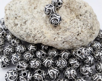 May include: A close-up of a pile of silver beads with a swirling design. The beads are arranged on a light gray rock.