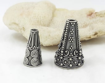 May include: Two silver cone-shaped beads with intricate designs. The larger bead has a pattern of dots and swirls, while the smaller bead has a pattern of spirals.