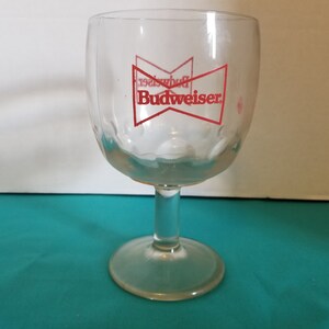 Puede incluir: Un vaso de cerveza Budweiser de vidrio transparente con un logotipo de Budweiser rojo y una corbata de lazo roja. El vaso tiene un cuenco redondeado y un tallo.