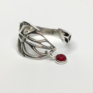 May include: A silver ring featuring a delicate, openwork design resembling a wing. A small red gemstone hangs from the ring.