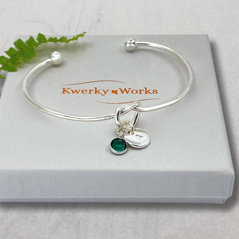 Personalised Silver Knot Bracelet with Initial and Birthstone Crystal: Meaningful Jewellery Gift for Women. This beautiful silver plated, open cuff, friendship knot bracelet makes a perfect gift for family or friend. It can be personalised with a birthstone crystal charm and a silver-plated initial letter charm of your choice. Here it is seen placed on top of the eco-friendly gift box that is included.