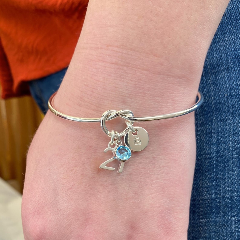 Picture of 21st birthday gift bracelet on a woman's arm. You can see how the charms hang beautifully from the knot of the bangle. The image shows the silver plated 21 charm, initial letter charm and choice of birthstone crystal colour to suit the birth month. This crystal is Aquamarine colour - for March birthstone
