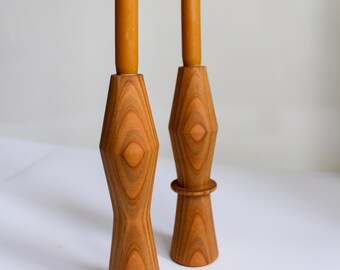 Puede incluir: Dos candelabros de madera con un diseño geométrico. Los candelabros están hechos de madera marrón claro y tienen un acabado natural. Cada candelabro tiene una sola vela amarilla alta.