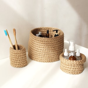 May include: Three woven jute baskets of varying sizes. The largest basket holds several bottles, the medium basket holds bottles, and the smallest basket holds toothbrushes. The baskets are a natural tan color and sit on a white surface.