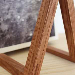 May include: A wooden picture frame stand with a triangular shape. The stand is made of dark brown wood with a natural wood grain. The stand is sitting on a light wood surface.