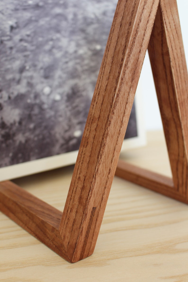 May include: A wooden picture frame stand with a triangular shape. The stand is made of dark brown wood with a natural wood grain. The stand is sitting on a light wood surface.