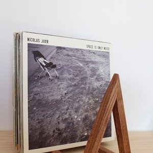May include: A wooden record stand with a triangular shape, holding a stack of vinyl records. The record on top is a black and white photo of a baby carriage on a dirt road. The album cover reads "Nicolas Jaar" and "Space is Only Noise".