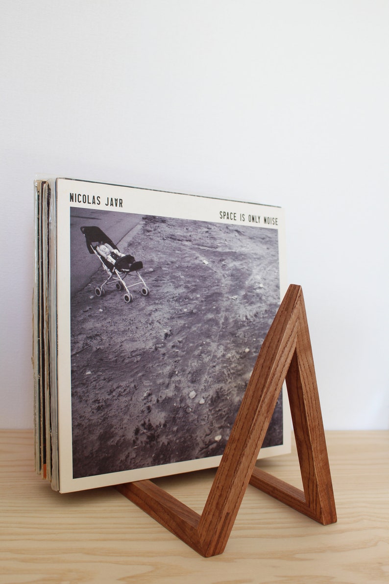 May include: A wooden record stand with a triangular shape, holding a stack of vinyl records. The record on top is a black and white photo of a baby carriage on a dirt road. The album cover reads "Nicolas Jaar" and "Space is Only Noise".