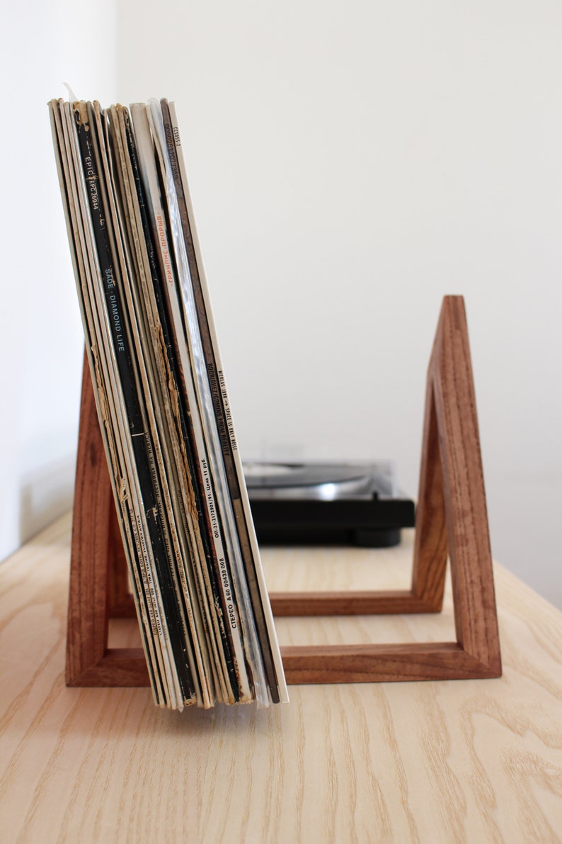 May include: A wooden record stand with a stack of vinyl records leaning against it. The stand is made of dark wood and has a simple, modern design.