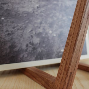 May include: A wooden picture stand with a natural wood finish. The stand is made of two pieces of wood that are joined together at a right angle. The stand is designed to hold a picture or photograph upright.