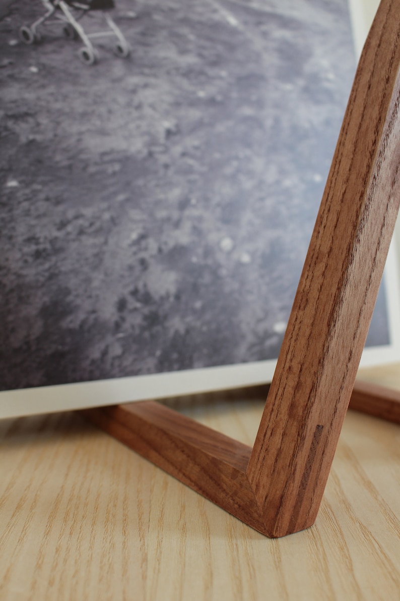 May include: A wooden picture stand with a natural wood finish. The stand is made of two pieces of wood that are joined together at a right angle. The stand is designed to hold a picture or photograph upright.