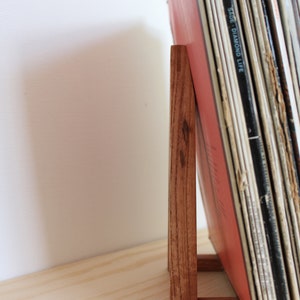 May include: A wooden record stand with a stack of vinyl records leaning against it. The stand is made of dark wood and has a simple design.