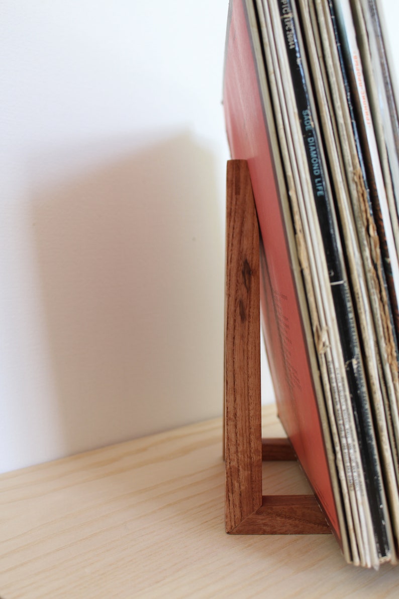 May include: A wooden record stand with a stack of vinyl records leaning against it. The stand is made of dark wood and has a simple design.