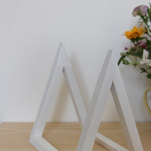 May include: Two white wooden triangle shelves, standing upright on a light wood surface. The shelves are made of simple, clean lines and are perfect for displaying small items.