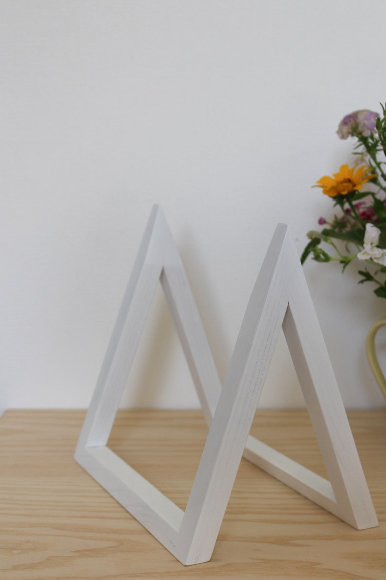 May include: Two white wooden triangle shelves, standing upright on a light wood surface. The shelves are made of simple, clean lines and are perfect for displaying small items.
