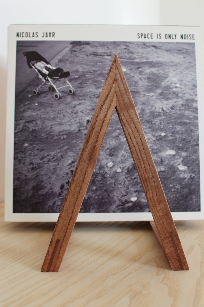 May include: A wooden triangle record stand with a record leaning against it. The record cover features a black and white photo of a baby in a stroller. The record title is "Space is Only Noise" by Nicolas Jaar.