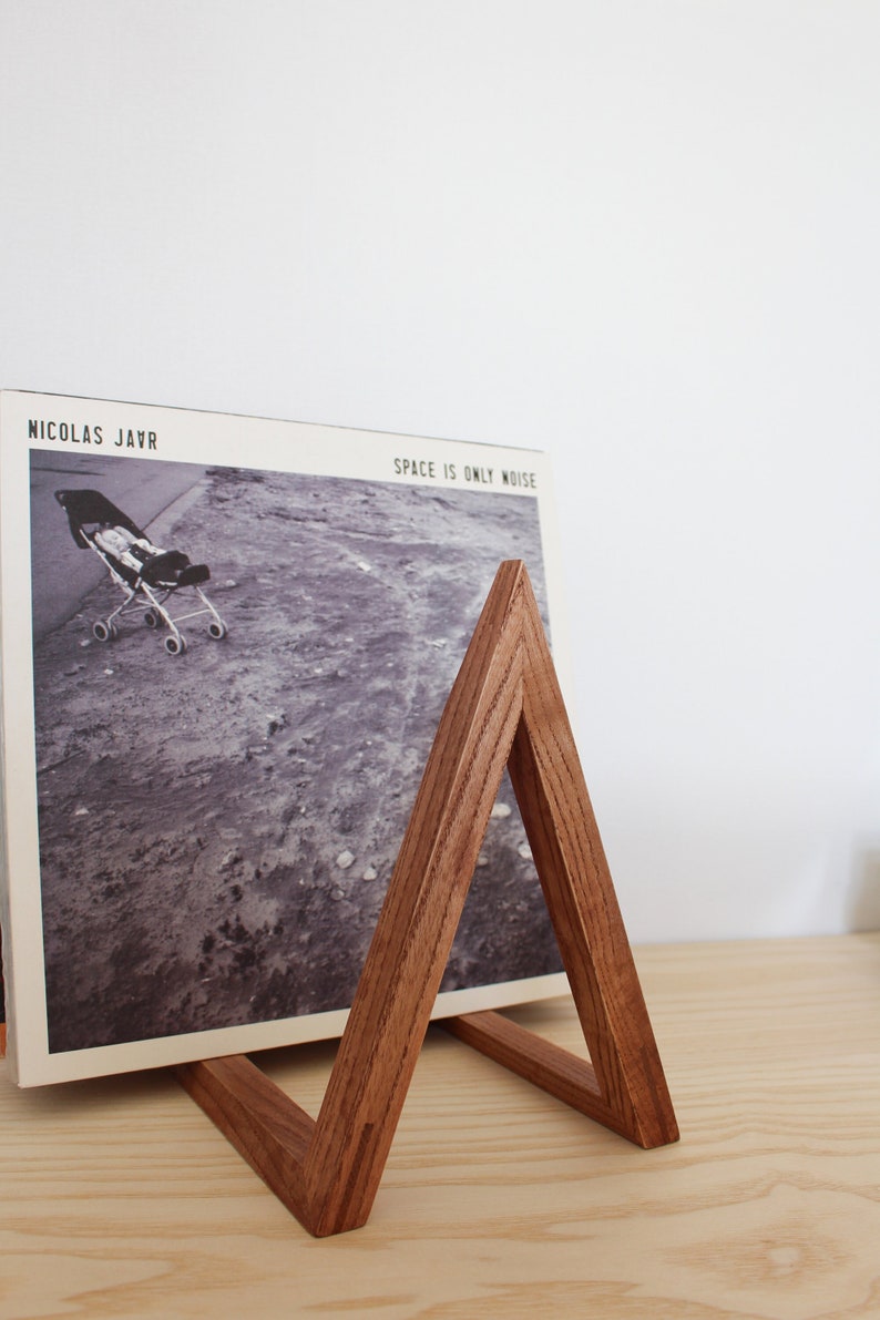 May include: A wooden record stand in the shape of a triangle, holding a vinyl record with the album title "Space is Only Noise" by Nicolas Jaar.