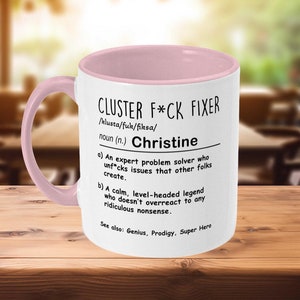 May include: White ceramic mug with a pink rim. The mug has a black and white design that reads "CLUSTER F*CK FIXER /klusta/fuk/fiksa/ noun (n.) Christine a) An expert problem solver who unf*cks issues that other folks create. b) A calm, level-headed legend who doesn't overreact to any ridiculous nonsense. See also: Genius, Prodigy, Super Hero"