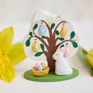 Puede incluir: Adorno de Pascua de madera con un árbol con adornos de huevo colgantes en azul, amarillo y rosa. Un conejito blanco está sentado junto a una cesta de huevos sobre una base verde. Una cinta crema está unida a la parte superior.