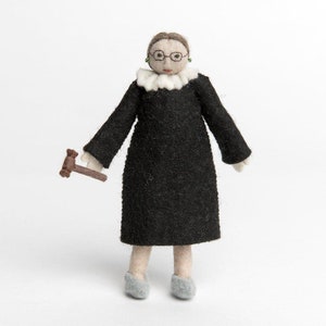 May include: A felt doll of a woman wearing a black robe and a white collar, holding a brown gavel. She has brown hair, glasses, and green earrings.