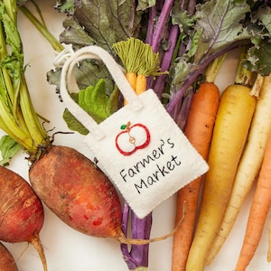 May include: A white felt tote bag with the words "Farmer's Market" and an apple design, filled with miniature felt carrots and green leaves, rests among colorful carrots, beets, and leafy greens.