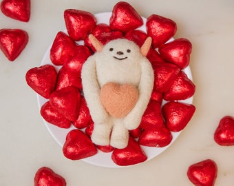 May include: A white felt creature with a pink heart in its arms, surrounded by red heart-shaped chocolates.