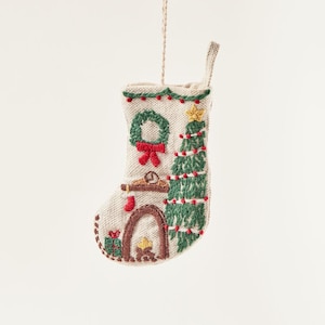 Puede incluir: Un adorno de calcetín de Navidad beige con detalles bordados, incluyendo una chimenea, un árbol de Navidad y una corona.