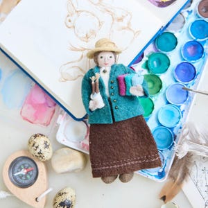 May include: A felt doll dressed in a teal jacket, brown skirt, and straw hat, holding paintbrushes and books. The doll is next to a watercolor palette, a compass, and speckled eggs. A sketchbook with a pencil drawing of a rabbit is in the background.