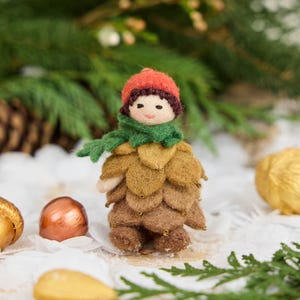 May include: A small felted figurine featuring a round face with dark hair, wearing a bright orange cap and a green scarf. The body is constructed from layered, overlapping felt petals in shades of golden yellow and brown, accented with tiny beads.